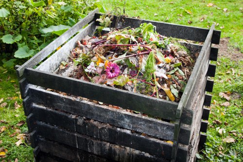 A black wooden compost bin situated outdoors on a grassy area with scattered fallen leaves, filled with a mixture of organic waste including decayed plant matter, vegetable scraps, and some colorful flowers. The compost is slightly overflowing and appears moist, with visible textures of decomposed materials. In the background, there is green foliage and a paved pathway, indicating a garden or backyard setting typical of residential rubbish removal. The image conveys organic waste collection, relevant to services offered by Commercial Waste Egham for garden and green waste disposal in the local area. The compost bin's slatted design allows air circulation, and the scene is illuminated with natural daylight, highlighting the textures and colours of the organic materials and surrounding greenery.