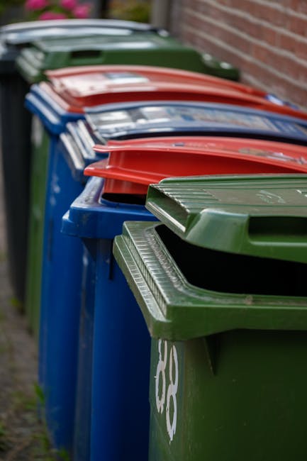 A row of multiple wheelie bins positioned along a pavement next to a brick wall, with the front bin being dark green, followed by blue, red, and green bins further down. Each bin has a hinged lid, wit