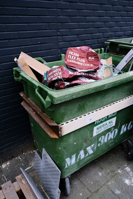 A row of multiple wheelie bins positioned along a pavement next to a brick wall, with the front bin being dark green, followed by blue, red, and green bins further down. Each bin has a hinged lid, wit